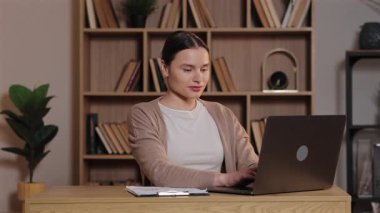 Masada oturup dizüstü bilgisayarla yazarken kameraya gülümseyen beyaz kadın. Evden çalışan kadın, sakin çalışma sahasında ahşap raflar ve bitkilerle ilerleme kaydetmekten memnun..