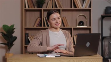Bilgisayardaki toplantı sırasında gülümseyen ve el sallayan beyaz bir kadın. Sanal sınıfa katılan kız öğrenci, kitaplarla dolu bir kitapla ev çalışma alanında ahşap masada oturuyor..
