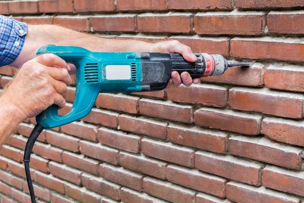 Braços segurando máquina de perfuração contra parede de tijolo — Fotografia de Stock