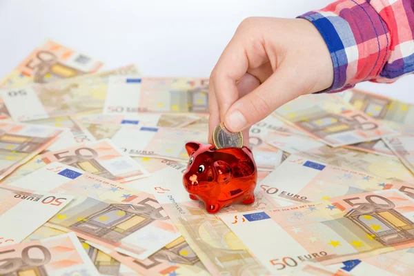 Mano poniendo moneda de euro en alcancía roja en billetes de euro — Foto de Stock