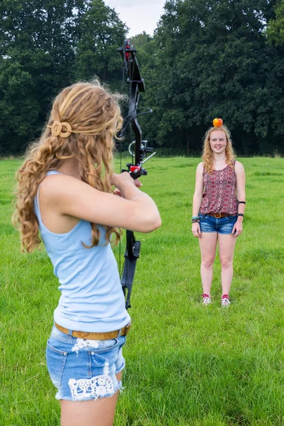 Girl aiming arrow of compound bow at apple on head of woman — Stock Photo, Image