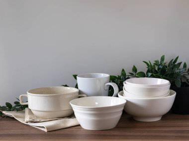 Yeşil yapraklı ahşap masaya dizilmiş beyaz seramik kaseler, fincanlar ve tabaklar. Temiz minimalist mutfak sofra takımı koleksiyonu sahnesi.