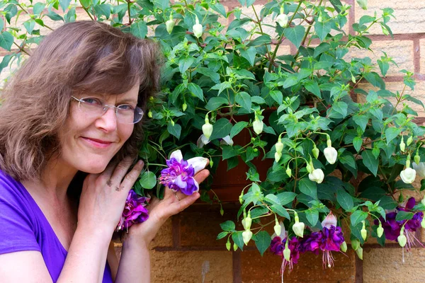 A beautiful mature woman is pictured enjoying the scent, size and majesty of Deep Purple fuchsia blooms.