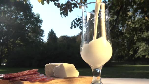 Verter la leche en un vaso en la mesa de picnic con queso y rebanadas de salchicha, de cerca — Vídeo de stock