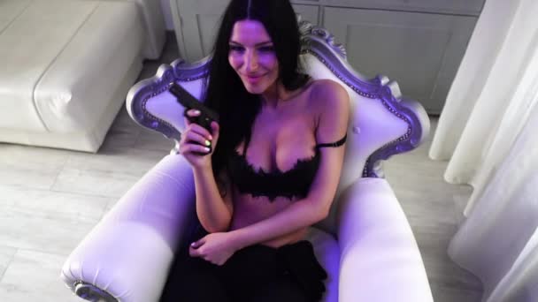 Соблазнительная молодая женщина в лифчике с пистолетом в руке сидит в роскошном кресле — стоковое видео