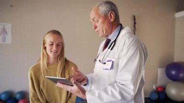 Erkek bir kıdemli doktorun rehabilitasyon merkezinde genç bir kadın hastayla kayıtları tartışırken görüntüsü.