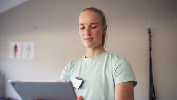 Porträt einer lächelnden blonden jungen Physiotherapeutin mit digitalem Tablet in der Hand in der Klinik — Stockvideo