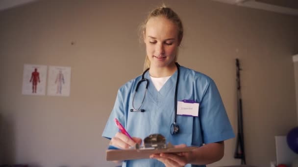 Zelfverzekerde vrouwelijke fysiotherapeut glimlacht en kijkt naar de camera tijdens het schrijven van het medisch rapport op klembord in de kliniek — Stockvideo