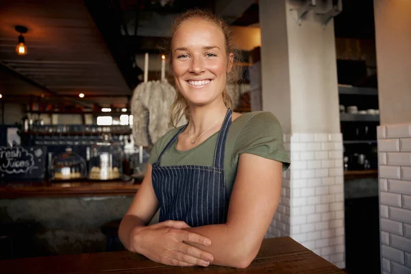 Vrolijke jonge vrouwelijke serveerster draagt schort leunend op tafel in cafe — Stockfoto