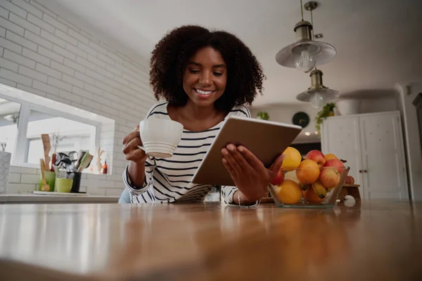 Усміхнена африканська жінка стоїть на кухні, спираючись на лічильник, використовуючи цифровий планшет під час пиття кави — стокове фото