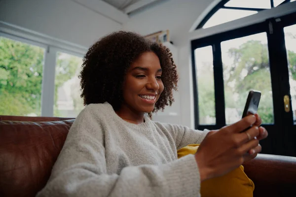 かなり黒の女性はソファに座っているスマートフォンを使用して家で時間を過ごす — ストック写真