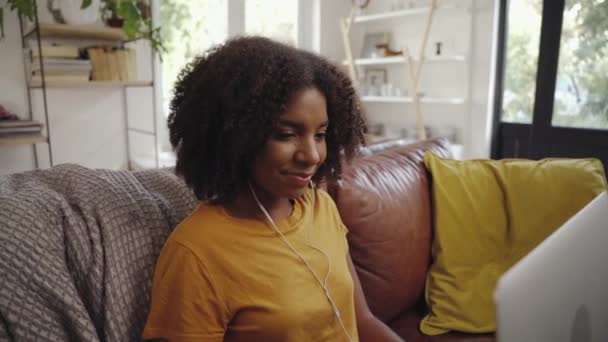 幸せな若いアフリカの女性ですリビングルームに座っている彼女の耳の中にイヤホンを使用してノートパソコン — ストック動画