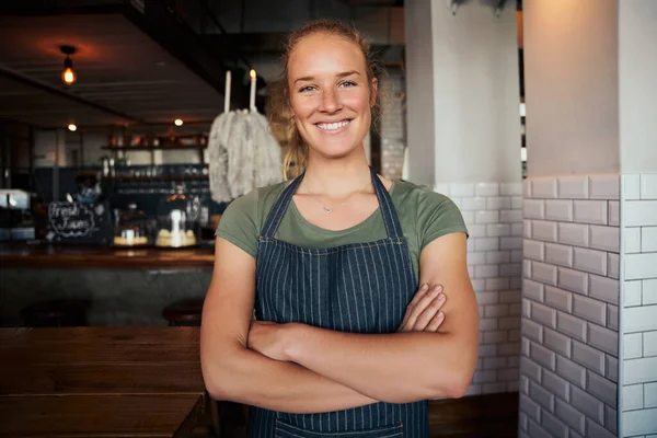 Portret van mooie jonge serveerster met armen gekruist in café en met schort aan — Stockfoto