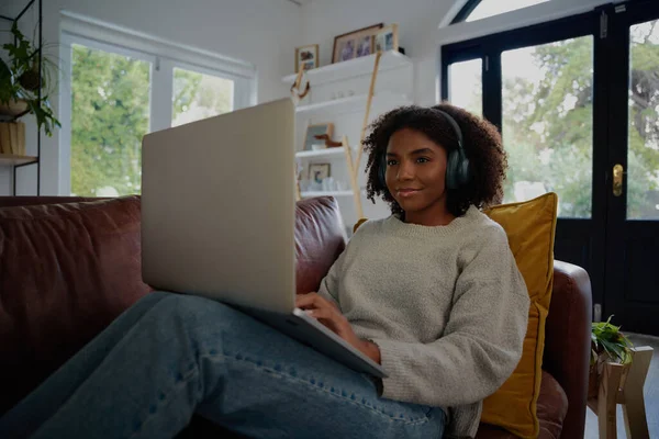 Retrato de mulher africana sentado usando laptop assistindo vídeo com fones de ouvido no sofá — Fotografia de Stock