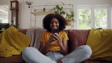 Gülümseyen, rahat Afro-Amerikan kadın koltukta oturuyor. Akıllı telefonu tut ve oturma odasında sosyal medya videosu izle.