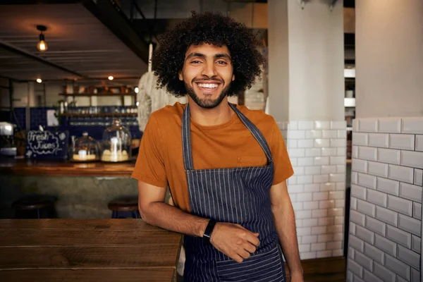 Portret van lachende jonge mannelijke ober met schort leunend op tafel staande kijkend naar camera in café — Stockfoto