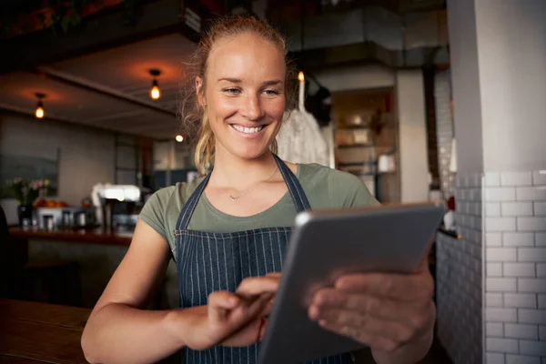Gelukkig vrouwelijke serveerster in cafe dragen schort met behulp van digitale tablet om online bestellingen te controleren — Stockfoto