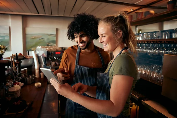 Alegre camarero masculino y femenino usando delantal usando tableta digital de pie detrás del mostrador en la cafetería — Foto de Stock
