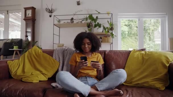 Zoom en la toma de una joven africana sentada en el sofá con las piernas cruzadas haciendo el pago en línea con tarjeta de crédito y teléfono inteligente — Vídeos de Stock