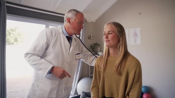 Positiv senior manlig sjukgymnast läkare kontrollerar den leende unga kvinnliga patienten lungor med stetoskop på kliniken — Stockvideo