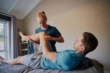 Genç adam hastane yatağında genç kadın fizyoterapiste eklem ağrısı pozisyonu gösteriyor.