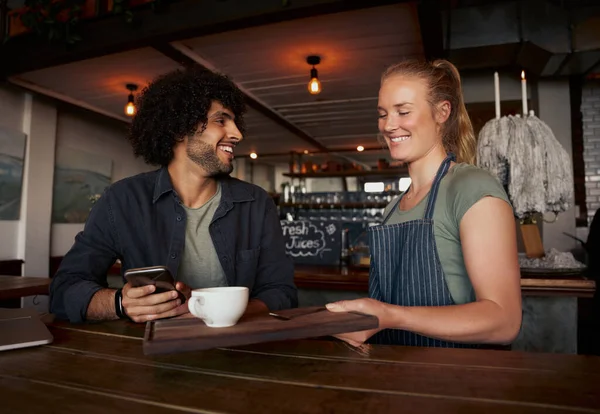 Garçonete feminina alegre servindo café para jovem cliente masculino com cabelo encaracolado usando smartphone no café moderno — Fotografia de Stock