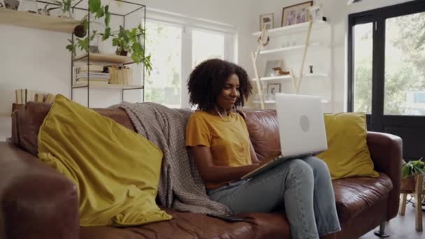 Retrato de jovem afro-americana sorridente com fones de ouvido digitando no laptop enquanto estava sentado no sofá na sala de estar — Vídeo de Stock