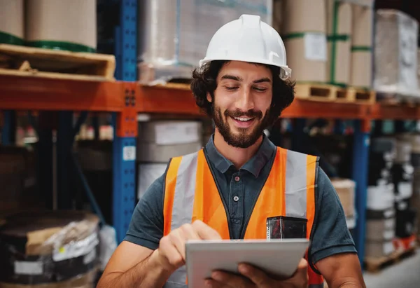 Jovem trabalhador sorridente caucasiano alegre com capacete na cabeça usando tablet para o trabalho, enquanto em pé no armazém contra mercadorias — Fotografia de Stock
