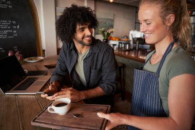 Beautiful young waitress serving man with curly hair with coffee on table clipart