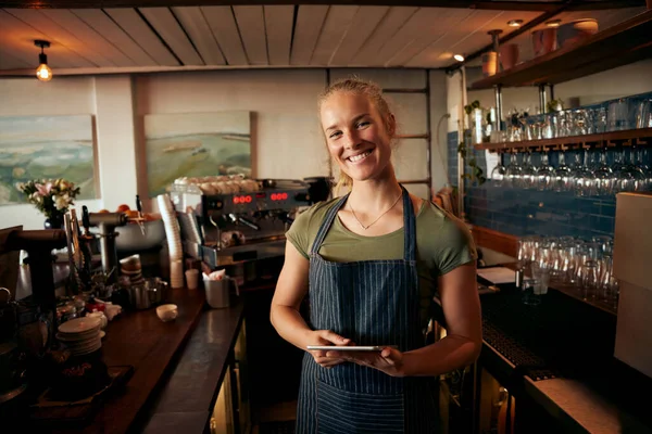 Portret van een mooie jonge vrouwelijke caféhouder die achter de toonbank staat en een schort draagt met digitale tablet — Stockfoto