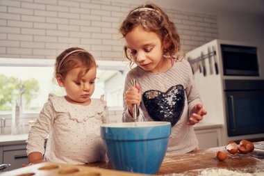 Evdeki mutfak tezgahında hamuru yumurtalarla hazırlarken küçük kızlar kasede el mikserini kullanıyorlar.