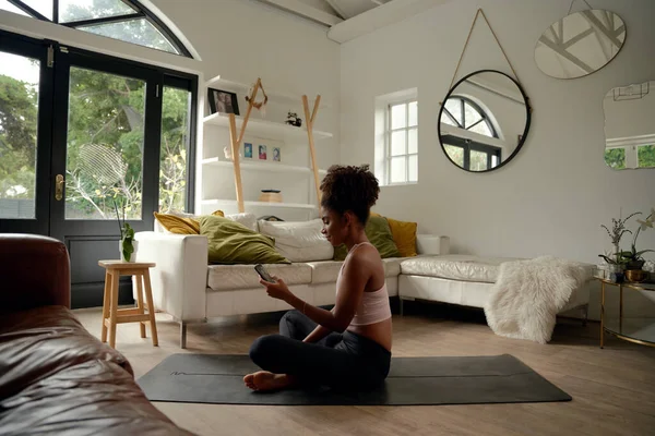 Молода африканська жінка використовує мобільний телефон, сидячи на йога мат вдома — стокове фото