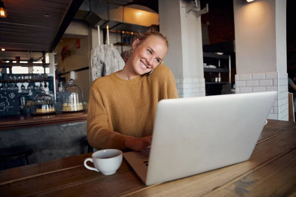 Χαρούμενη νεαρή γυναίκα επιχειρηματίας κάθεται στο σύγχρονο καφέ μιλώντας μέσω τηλεφώνου και χρησιμοποιώντας το laptop — Φωτογραφία Αρχείου
