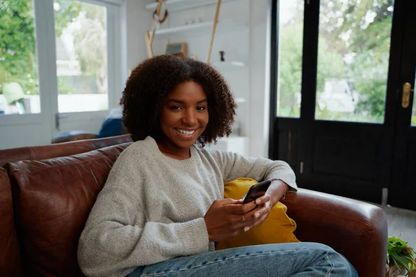 カメラを見てソファに座って携帯電話を保持している陽気なアフリカの女性の肖像画 — ストック写真