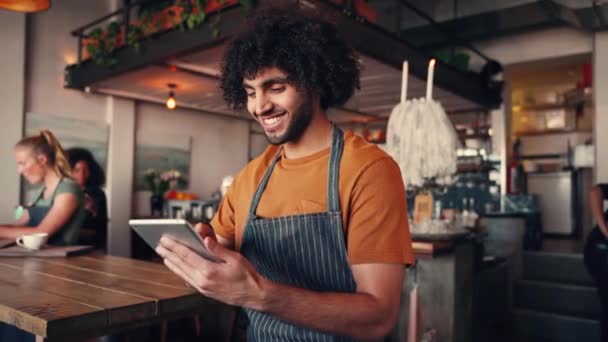 コーヒーショップでデジタルタブレットを使用してエプロンを身に着けている成功した笑顔の若者のショット — ストック動画