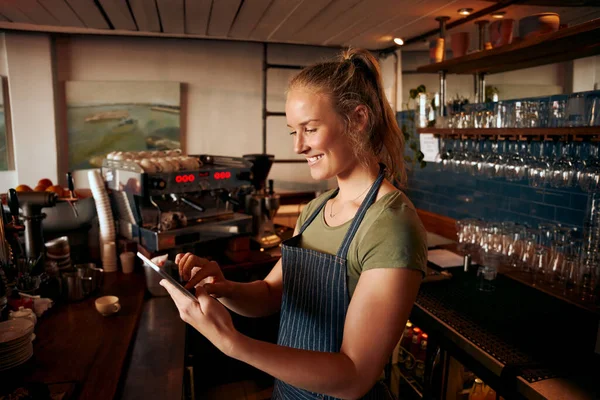 Ung kvinnlig personal som bär förkläde och arbetar med en digital surfplatta vid caféets disk — Stockfoto