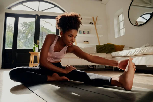 Тонка спортсменка в спортивному одязі розігріває м'язи перед тренуванням, роблячи вправи для розтягування, сидячи на підлозі вдома — стокове фото