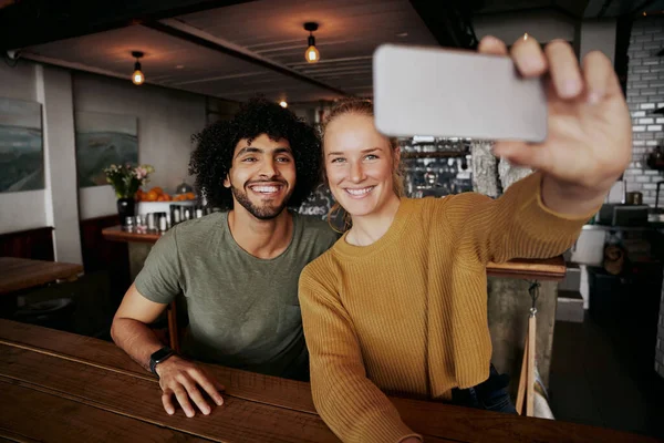 Mujer joven sonriente sentada con un amigo varón tomando selfie en la cafetería usando el teléfono móvil — Foto de Stock