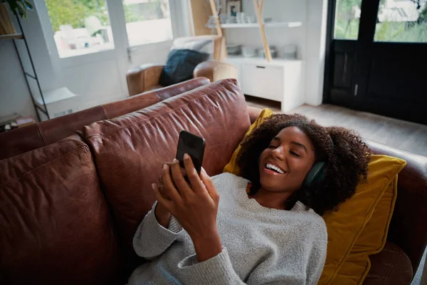 家でスマートフォンを使用してソファの上に横たわるおもちゃの笑顔を持つ女性ブルートゥースワイヤレスヘッドフォンで音楽を聴く — ストック写真