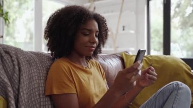 Elinde kredi kartı ve akıllı telefon olan gülümseyen Afrikalı bir kadın evdeki kanepede oturuyor.