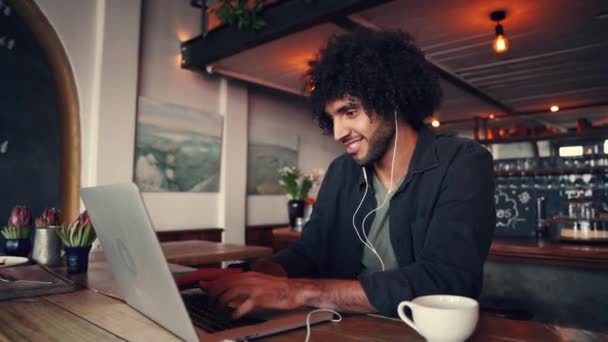 Glücklicher junger Mann mit Kopfhörer in den Ohren tippt auf Laptop, während er am Tisch im Café sitzt — Stockvideo