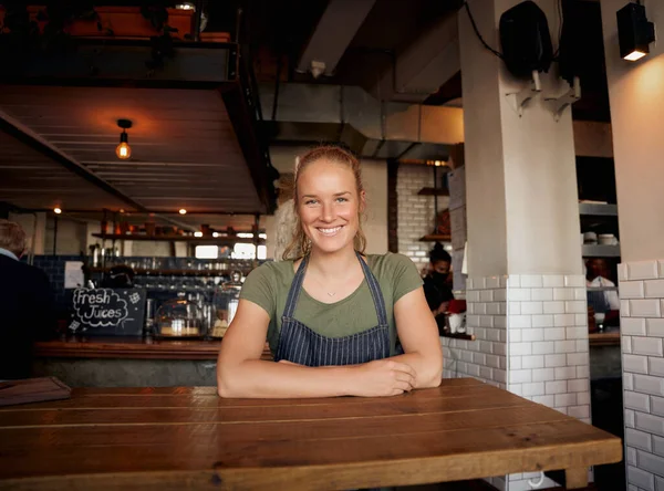 Fröhliche junge Café-Inhaberin mit Schürze lächelt in die Kamera und lehnt sich auf den Tisch — Stockfoto