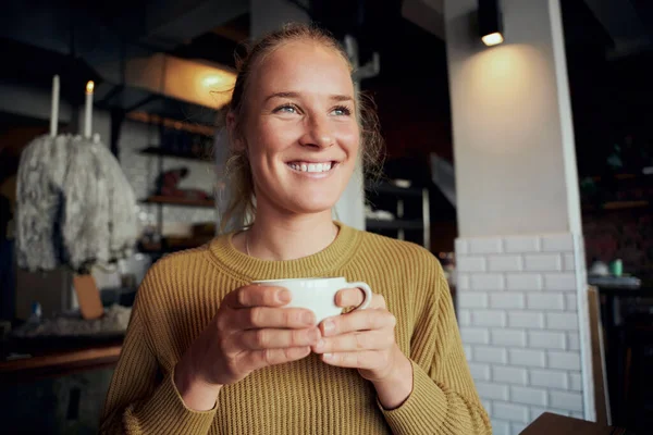 Mooie jonge vrouw met koffiebeker in cafetaria glimlachend en wegkijkend — Stockfoto