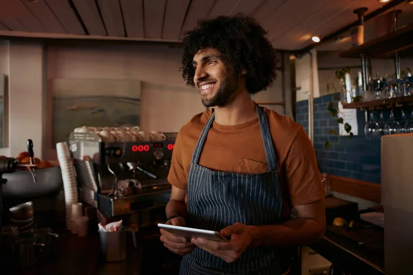 Successful small male business owner wearing apron using digital tablet and looking away
