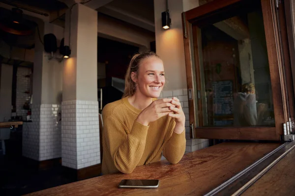Wesoła kobieta pijąca kawę w kawiarni odwracając wzrok telefonem na stole — Zdjęcie stockowe