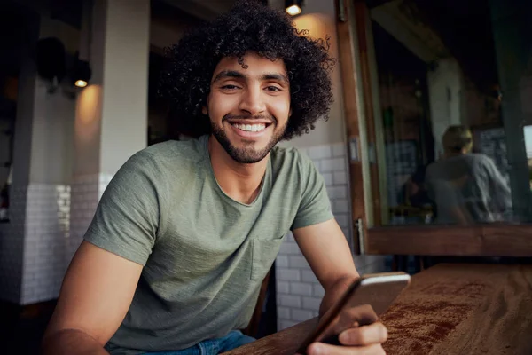 Retrato del hombre guapo sonriente sentado en la cafetería sosteniendo el teléfono mirando a la cámara — Foto de Stock