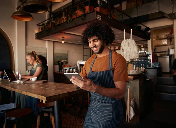 Mannelijke ober met krullend haar werken in cafe dragen schort met behulp van digitale tablet met vrouwelijke werken op de achtergrond op laptop — Stockfoto