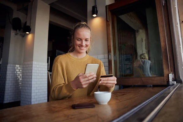 Jovem mulher fazendo pagamento on-line usando cartão e smartphone enquanto está sentado e desfrutando de café no café — Fotografia de Stock