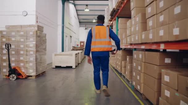 Follow shot rear view of male warehouse worker holding hardhat in hand walking down factory storage — Vídeo de Stock