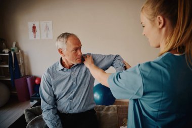 Genç bayan fizyoterapist hastane odasında yaşlı bir hastanın omuz ve dirseğini muayene ediyor.